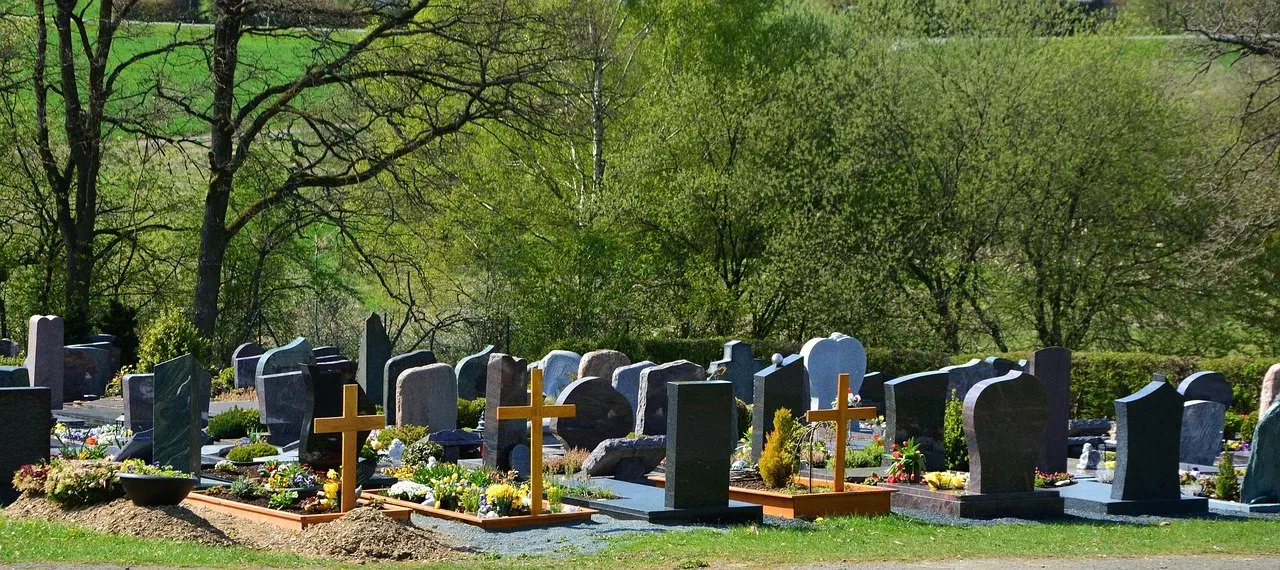 Friedhof mit Grabsteinen in mehreren Reihen, vorn frische Gräber mit hellen Holzkreuzen und Blumen, dahinter Bäume und Wiese