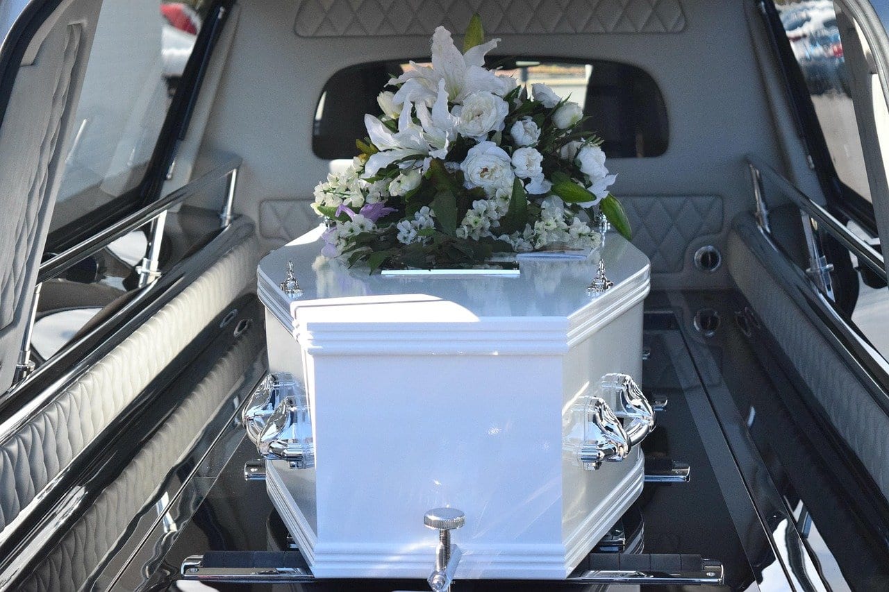 Weißer Sarg mit Gesteck aus weißen Rosen und Lilien im Laderaum eines Bestattungswagens mit geöffneter Heckklappe