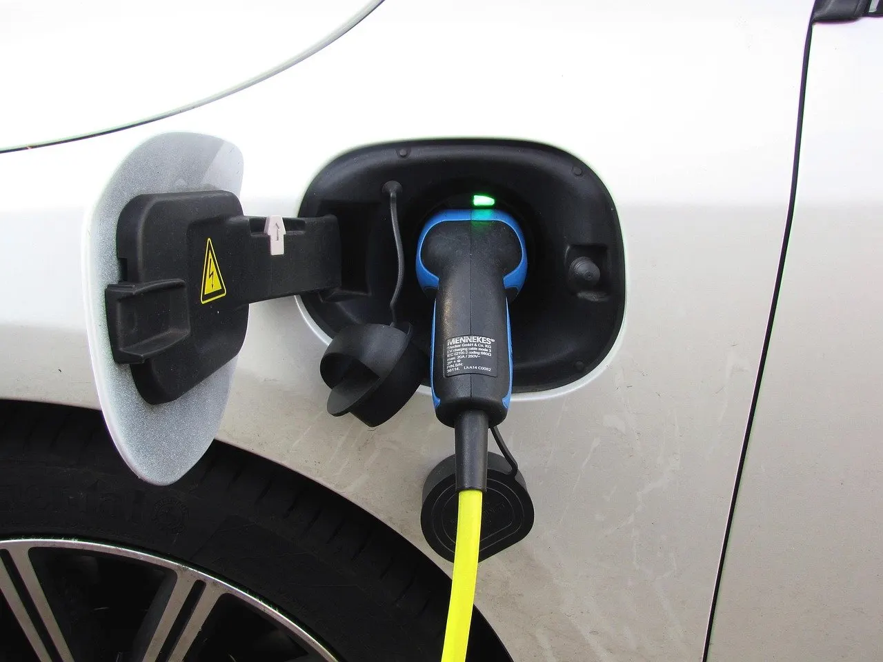Ladestecker mit gelbem Kabel steckt in der geöffneten Ladebuchse eines weißen Elektroautos, am Stecker leuchtet eine grüne Lampe