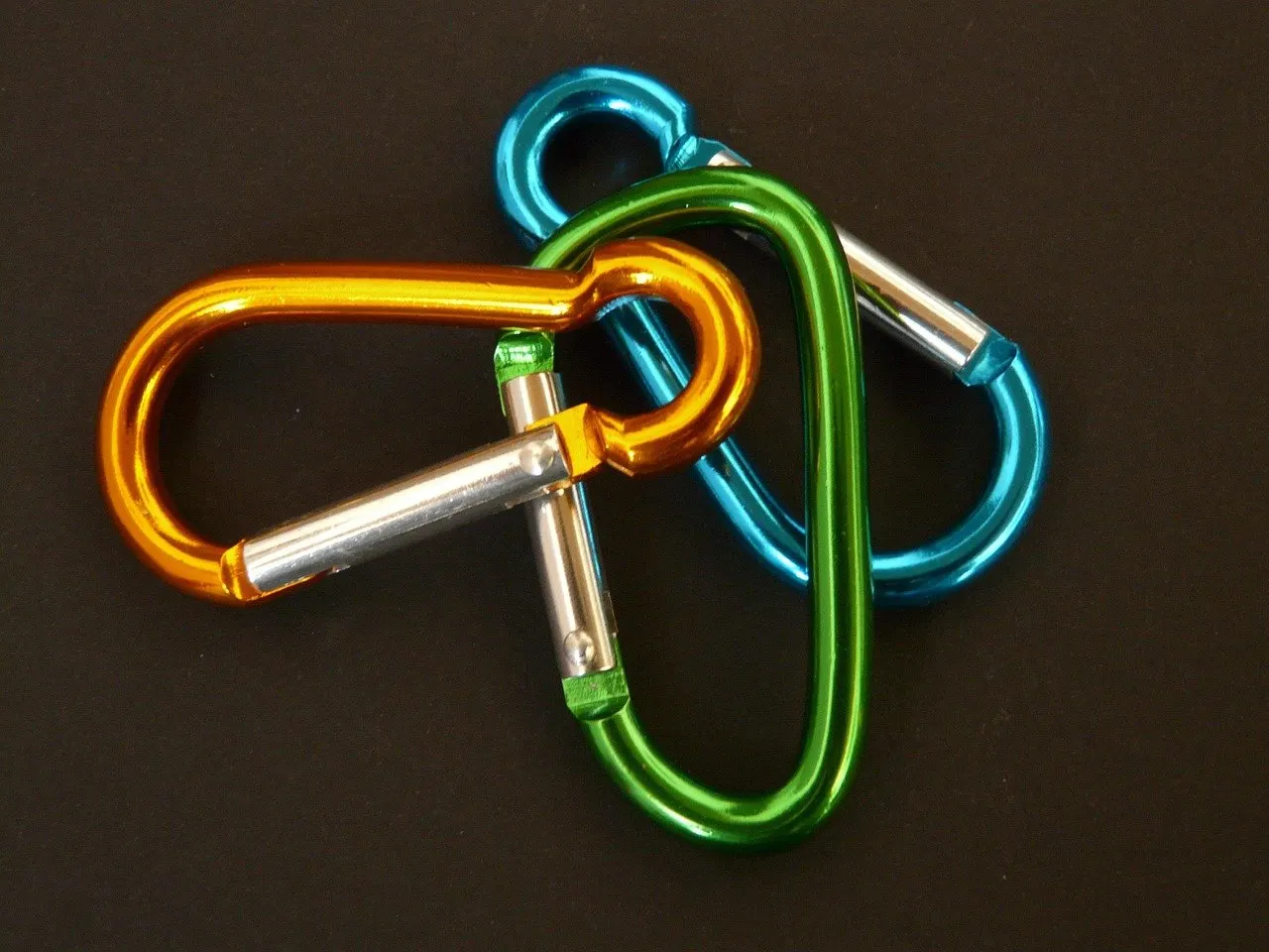 Drei farbige Karabinerhaken aus Aluminium in Orange, Grün und Blau mit silbernen Schnappern auf dunklem Untergrund