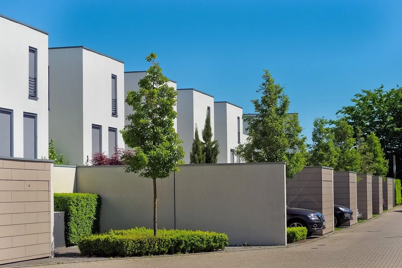 Reihe moderner weißer Einfamilienhäuser mit Flachdach hinter Gartenmauern, davor junge Bäume, Hecken, geparkte Autos und eine gepflasterte Straße