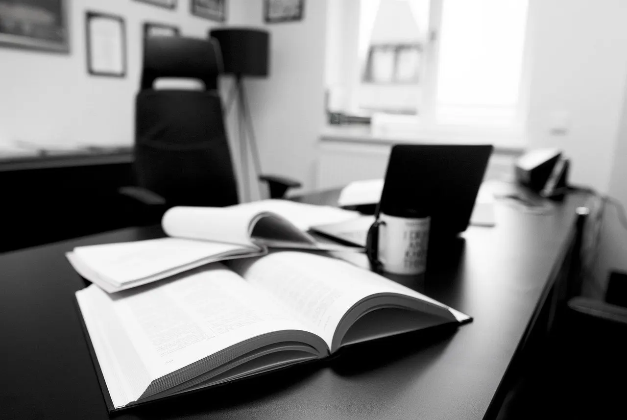 Schwarz-Weiß-Aufnahme: Schreibtisch in einer Kanzlei mit zwei aufgeschlagenen Büchern, Laptop und Tasse, dahinter ein leerer Bürostuhl und ein Fenster