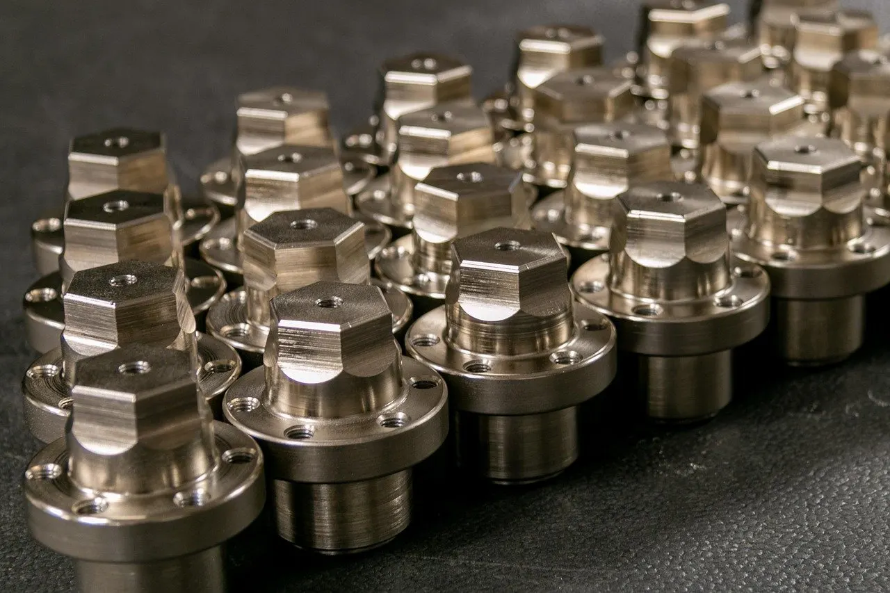 Viele gedrehte Metallteile mit Sechskantkopf und Flansch stehen in Reihen auf dunklem Untergrund, ihre Oberflächen glänzen metallisch