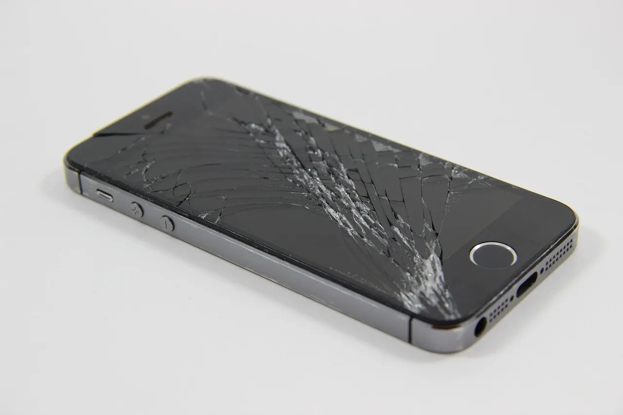 Dunkelgraues Smartphone mit stark zersplittertem Displayglas liegt schräg auf einer hellen, weißen Unterlage