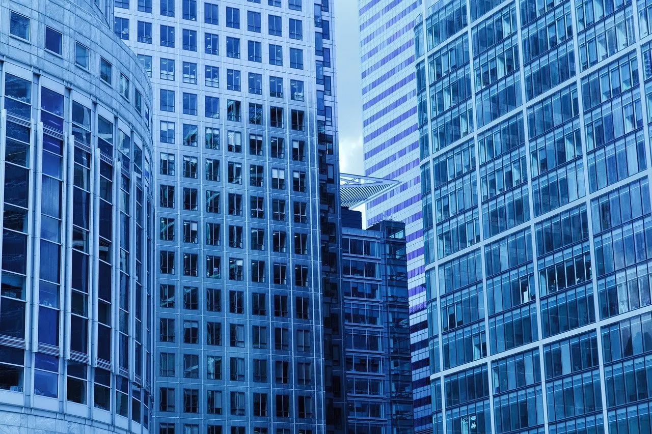 Mehrere hohe Bürogebäude mit Glas- und Rasterfassaden, von unten fotografiert, dazwischen ein schmaler Streifen bewölkter Himmel