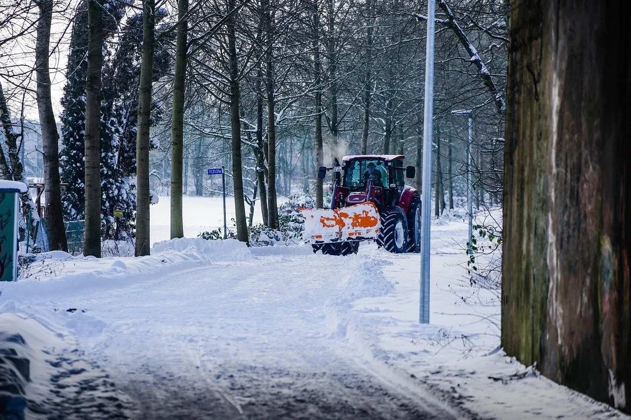 Roter Traktor mit orangefarbenem Räumschild schiebt Schnee auf einem verschneiten Weg zwischen kahlen Bäumen und Laternen